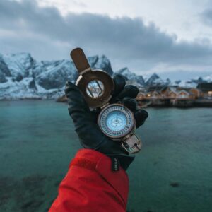 A gloved hand holds a compass in Reine, Lofoten Islands, with a stunning fjord backdrop.